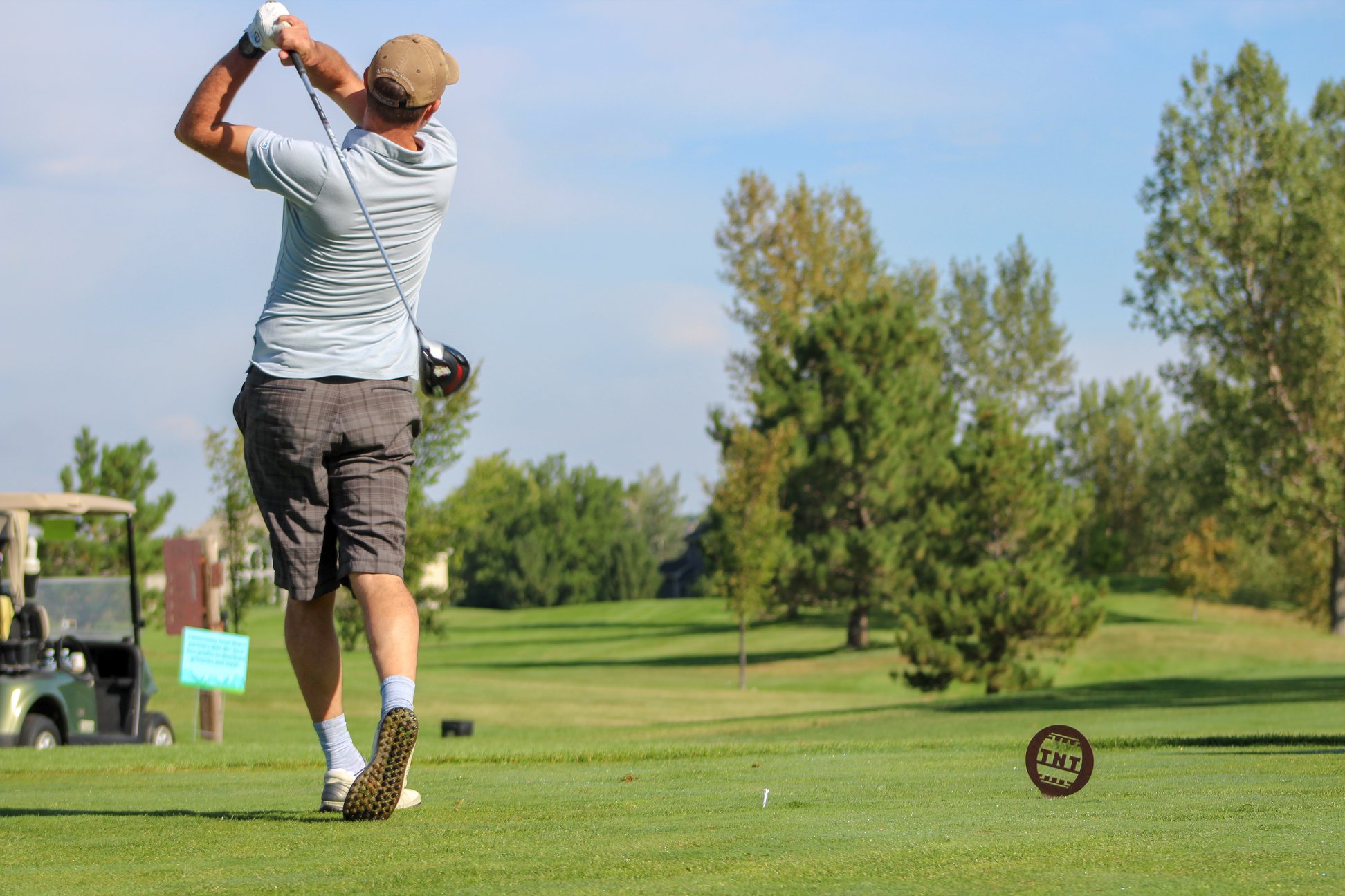 A man swings a golf club, with trees and a golf cart in the background.