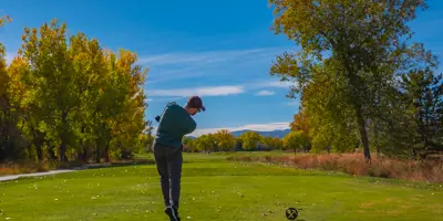 A man swings a golf club, with trees and a blue sky in the background.