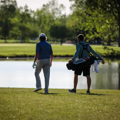 Two people carrying golf equipment stand next to a pond, with trees in the background.