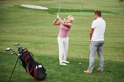 A woman swinging a golf club, with a man and a golf bag in the foreground.