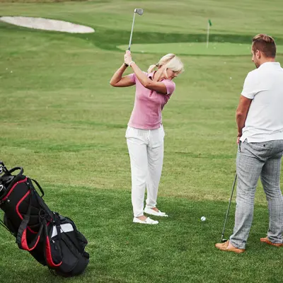 A woman swinging a golf club, with a man and a golf bag in the foreground.